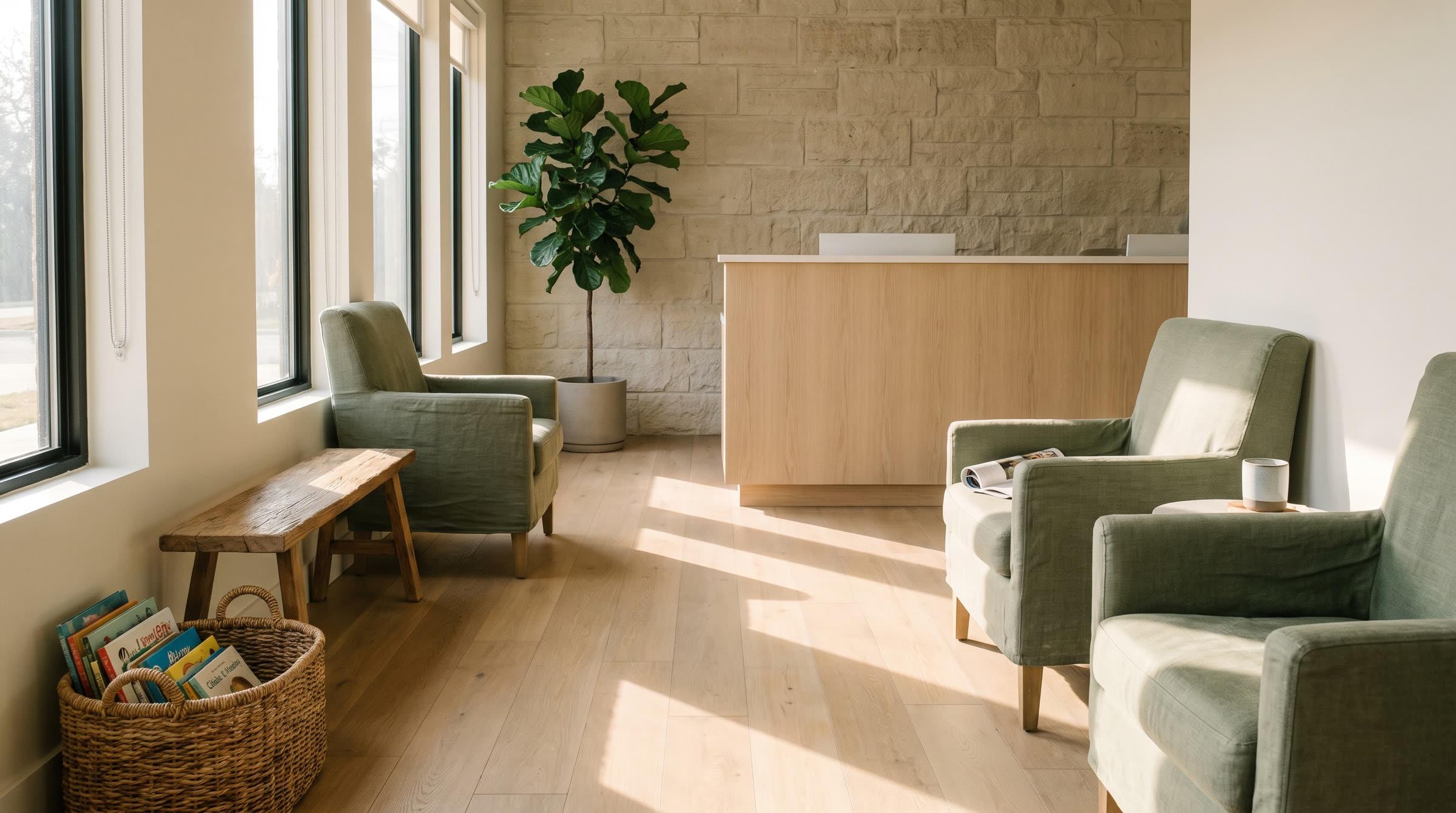 A sunlit waiting room with sage chairs, a fig tree, and a pale oak desk