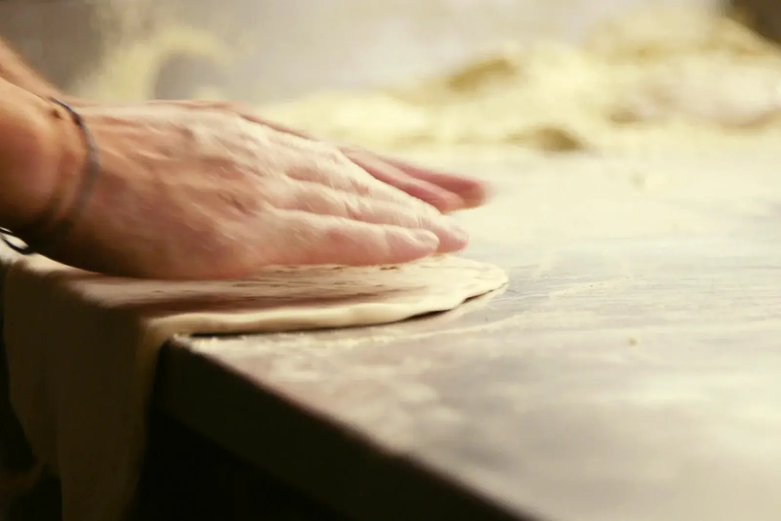 Hands pressing a round of pizza dough flat on a floured steel counter.