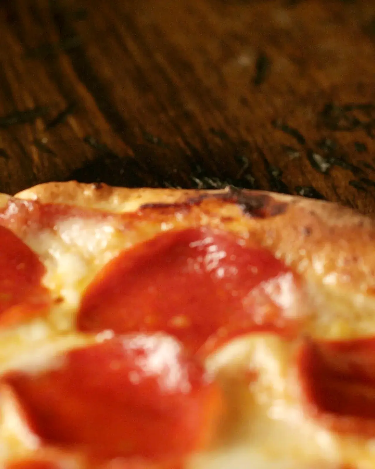 Close-up of crisp, cupped pepperoni and a golden, blistered crust on a dark wood table.