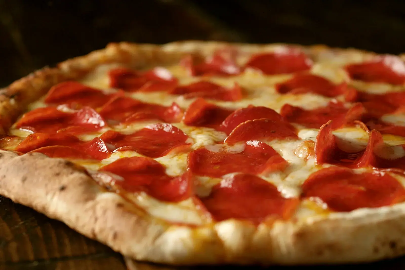 A whole pepperoni pizza with a puffy golden crust resting on a dark wood table.